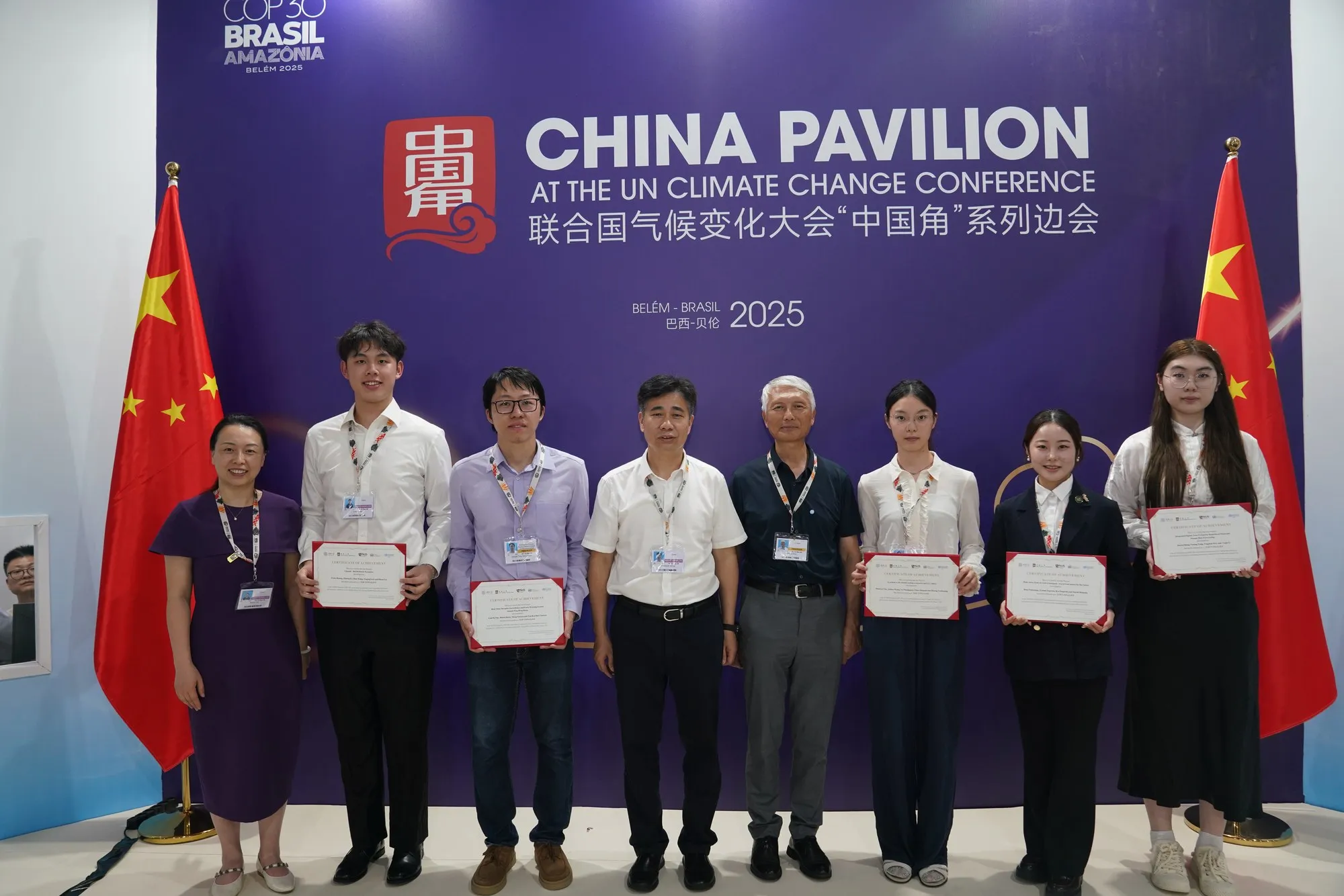 ベレンのCOP30で、中国パビリオンの背景の前に並ぶ8人。そのうち5人が証書を持っている。
