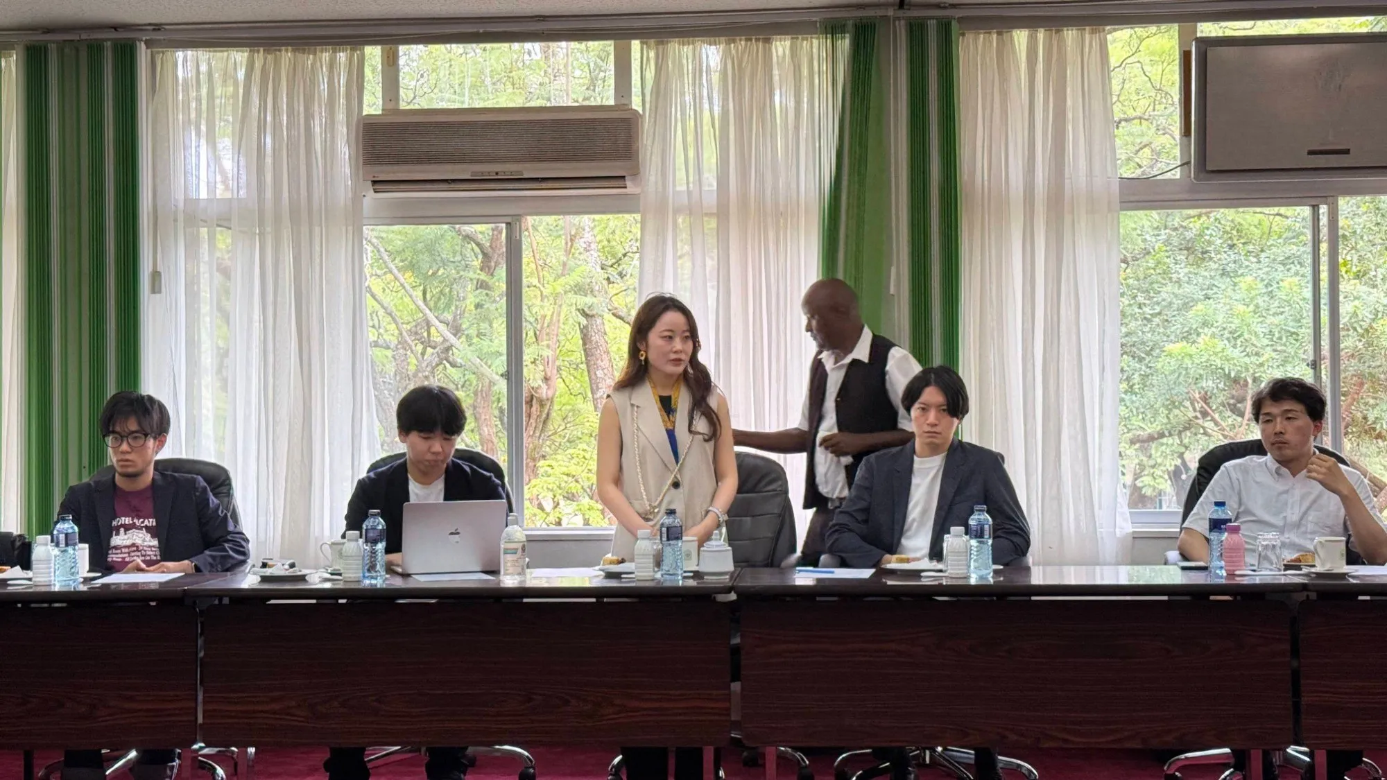 Programme participants seated along a table in a meeting room; one of them stands to speak.